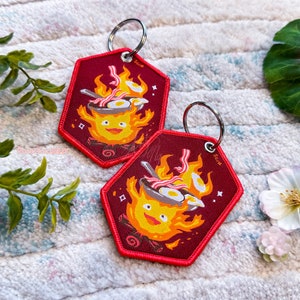 May include: Two red fabric keychains featuring a cartoon character resembling a fire spirit cooking bacon and eggs in a pan. The character has a yellow body and a smiling face. The keychains are on a white fluffy surface.