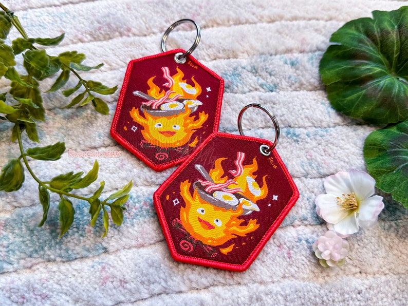 May include: Two red fabric keychains featuring a cartoon character resembling a fire spirit cooking bacon and eggs in a pan. The character has a yellow body and a smiling face. The keychains are on a white fluffy surface.
