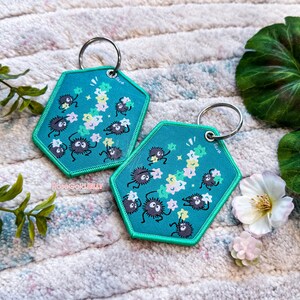 May include: Two teal blue hexagonal keychains with a white string of stars and black fuzzy creatures holding stars. The keychains are on a white and pink fluffy surface.