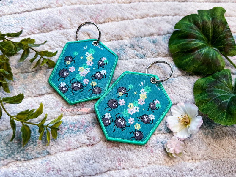 May include: Two teal blue hexagonal keychains with a white string of stars and black fuzzy creatures holding stars. The keychains are on a white and pink fluffy surface.