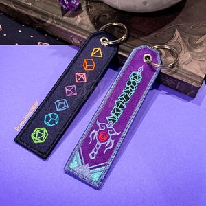 May include: Two keychains with embroidered designs. One keychain is black with a row of colorful dice. The other keychain is purple with a sword made of dice. Both keychains have silver key rings.
