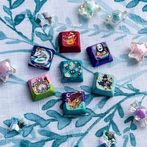 Puede incluir: Una colecci&oacute;n de siete teclas cuadradas para teclado mec&aacute;nico con coloridas ilustraciones inspiradas en el anime, que incluyen personajes como un duende del polvo, un gato-bus y un esp&iacute;ritu, acompa&ntilde;adas de varias cuentas transl&uacute;cidas con forma de estrella.