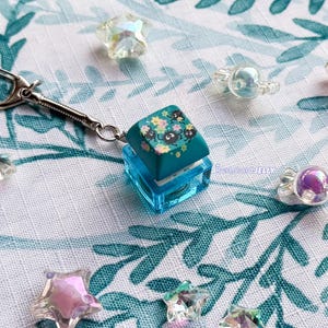 Puede incluir: Un llavero con una tecla de teclado mec&aacute;nico de color verde azulado con un estampado floral y peque&ntilde;os duendes del holl&iacute;n negros. La tecla est&aacute; montada sobre una base cuadrada azul transl&uacute;cida con un cierre de mosquet&oacute;n plateado y cuentas.