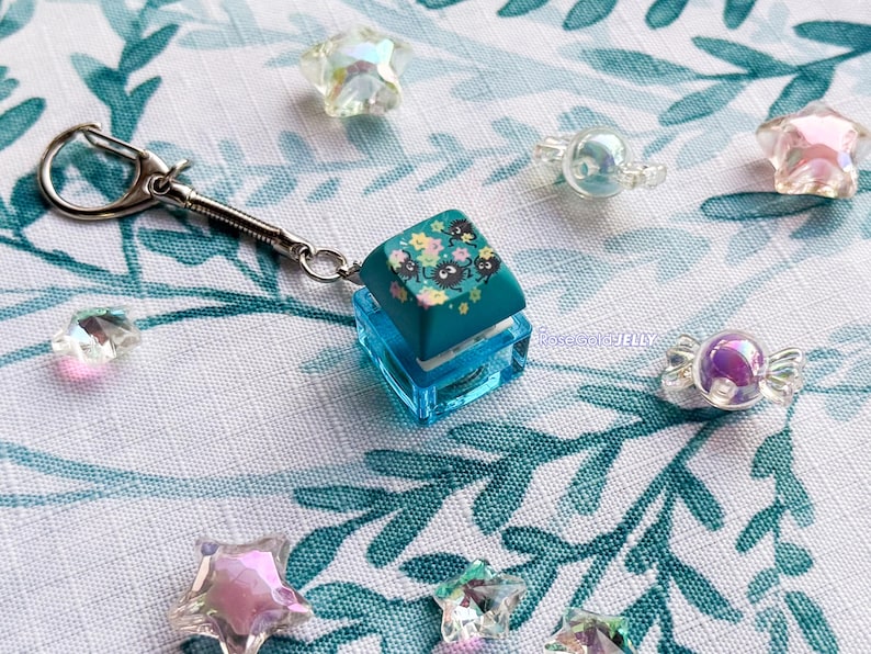 Puede incluir: Un llavero con una tecla de teclado mec&aacute;nico de color verde azulado con un estampado floral y peque&ntilde;os duendes del holl&iacute;n negros. La tecla est&aacute; montada sobre una base cuadrada azul transl&uacute;cida con un cierre de mosquet&oacute;n plateado y cuentas.