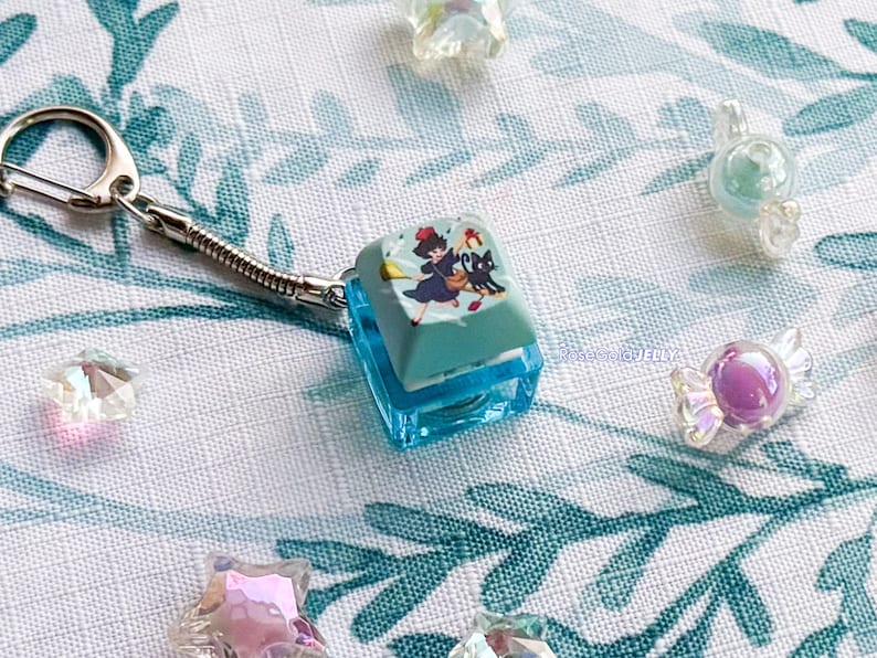 Puede incluir: Un llavero con una tecla de teclado mec&aacute;nico de color azul claro que presenta un gr&aacute;fico de un personaje con vestido oscuro y un gato negro sobre una escoba. La tecla est&aacute; unida a una anilla de metal plateado y rodeada de peque&ntilde;os dijes transl&uacute;cidos con forma de estrella y caramelo.