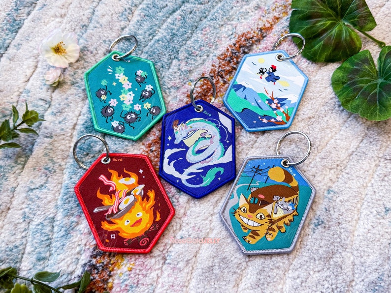 May include: Five colourful keychains featuring characters from popular animated films. The keychains are hexagonal shaped and feature embroidered designs. The keychains are on a white fluffy surface.