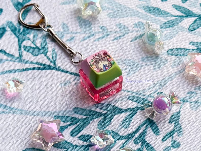 Puede incluir: Un llavero de tecla de teclado mec&aacute;nico con una parte superior en degradado morado y verde, con la ilustraci&oacute;n de una criatura gris y un peque&ntilde;o personaje azul. La base es una carcasa cuadrada de color rosa transl&uacute;cido unida a una anilla met&aacute;lica.