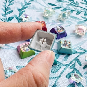 Puede incluir: Una tecla de teclado mec&aacute;nico con exterior azul claro e interior blanco con un v&aacute;stago en forma de cruz. La tecla est&aacute; rodeada por varias otras teclas coloridas con diversas ilustraciones de personajes y peque&ntilde;as cuentas transparentes en forma de estrella.
