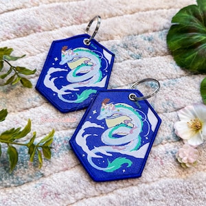 May include: Two blue hexagonal keyrings featuring a white dragon with a girl riding on its back. The dragon is surrounded by white clouds and stars against a blue background. The keyrings have silver key rings.