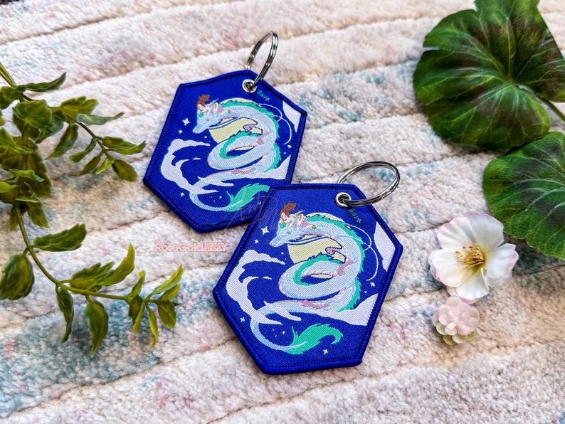 May include: Two blue hexagonal keyrings featuring a white dragon with a girl riding on its back. The dragon is surrounded by white clouds and stars against a blue background. The keyrings have silver key rings.