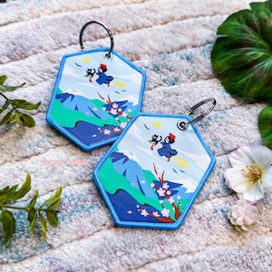 May include: Two blue hexagonal keyrings with embroidered images of a witch flying on a broomstick with a black cat. The keyrings feature a mountain landscape with cherry blossoms.