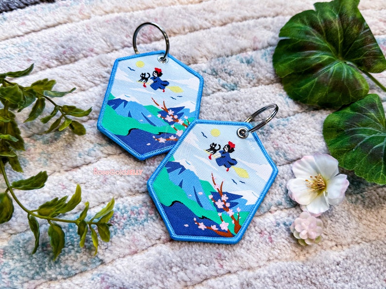 May include: Two blue hexagonal keyrings with embroidered images of a witch flying on a broomstick with a black cat. The keyrings feature a mountain landscape with cherry blossoms.