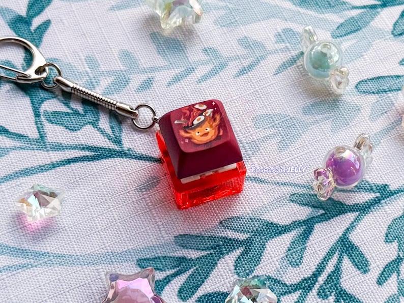 Puede incluir: Un llavero con una tecla de teclado mec&aacute;nico de color granate que presenta un gr&aacute;fico de una peque&ntilde;a criatura naranja cocinando huevos en una sart&eacute;n. La tecla est&aacute; unida a una anilla de metal plateado sobre una base de pl&aacute;stico rojo.