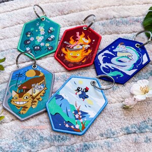 May include: Five colourful keychains featuring characters and scenes from the Studio Ghibli films. The keychains are made of fabric and have a hexagonal shape. The keychains feature characters such as Totoro, No Face, and Kiki.