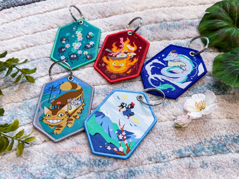 May include: Five colourful keychains featuring characters and scenes from the Studio Ghibli films. The keychains are made of fabric and have a hexagonal shape. The keychains feature characters such as Totoro, No Face, and Kiki.
