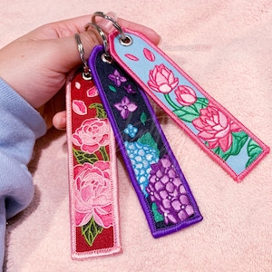 May include: Three embroidered keyrings with floral designs. One is red with pink roses, another is purple with hydrangeas, and the last is light blue with pink lotus flowers. Each keyring has a metal ring for keys.