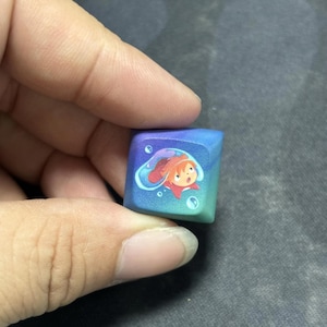 Puede incluir: Una tecla mec&aacute;nica cuadrada para teclado con un degradado de color que va del morado intenso al verde azulado. El centro de la tecla muestra un peque&ntilde;o personaje ilustrado con cabello naranja dentro de una burbuja de agua azul transl&uacute;cida.