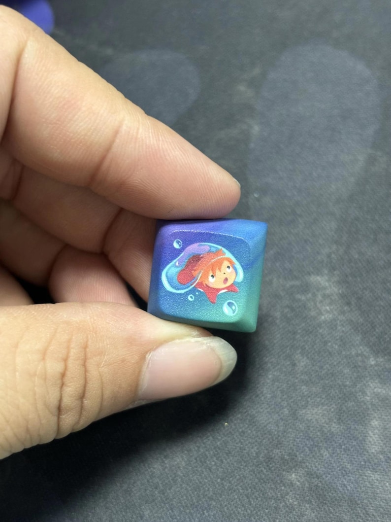 Puede incluir: Una tecla mec&aacute;nica cuadrada para teclado con un degradado de color que va del morado intenso al verde azulado. El centro de la tecla muestra un peque&ntilde;o personaje ilustrado con cabello naranja dentro de una burbuja de agua azul transl&uacute;cida.