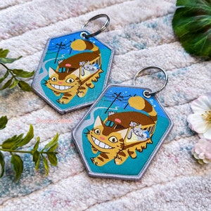 May include: Two keychains featuring a cartoon cat riding a bus with a blue background. The cat has a large smile and is brown and orange. The bus is brown and has a white roof. The keychains are silver and have a silver ring.
