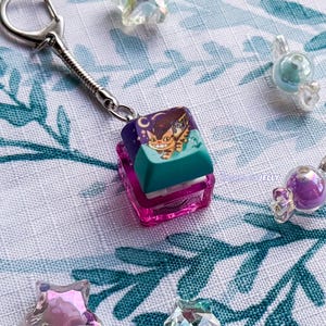 Puede incluir: Un llavero con una tecla de teclado mec&aacute;nico cuadrada con una base en degradado morado y verde azulado. La parte superior de la tecla muestra un gr&aacute;fico de un gato naranja sonriente con ojos grandes sobre un cielo morado oscuro con una luna.