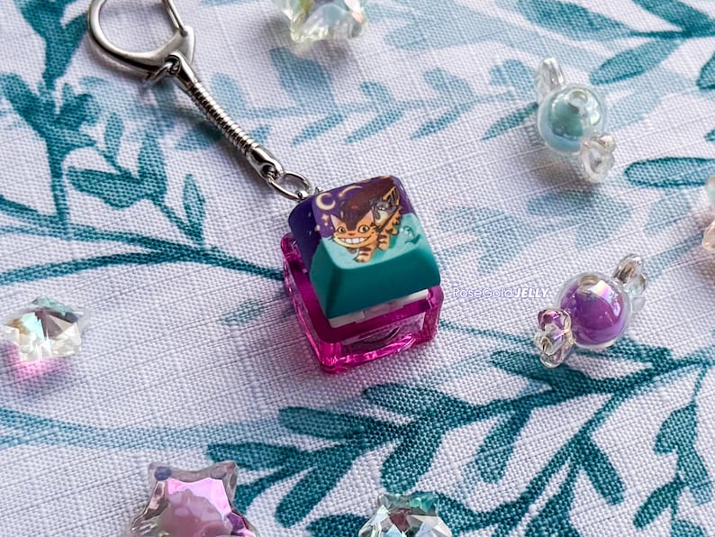 Puede incluir: Un llavero con una tecla de teclado mec&aacute;nico cuadrada con una base en degradado morado y verde azulado. La parte superior de la tecla muestra un gr&aacute;fico de un gato naranja sonriente con ojos grandes sobre un cielo morado oscuro con una luna.