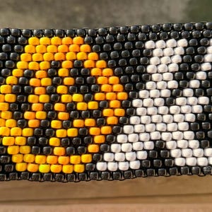 May include: A rectangular cuff bracelet made of black plastic pony beads. The design features the word "LUCK" spelled out in beads, with the "L" in blue, the "U" in orange, the "C" in white, and the "K" in green, all set against a black background.