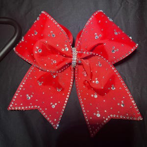 May include: A bright red cheerleading bow featuring a sparkling rhinestone design. The bow is decorated with many small rhinestones and a central rhinestone detail. The bow is made of a ribbed fabric.