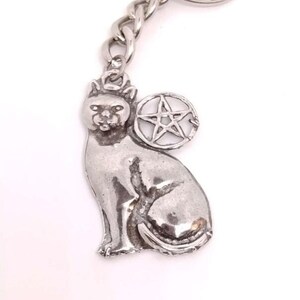 May include: Silver-toned cat keychain with a pentagram charm. The cat is in a seated position, with a detailed face and body. The keychain has a chain link and a circular key ring.