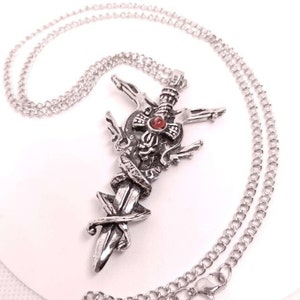 May include: A silver-colored pendant necklace featuring a sword design with a red gemstone at the center. The pendant is intricately detailed with a snake wrapped around the sword. The necklace has a delicate chain.