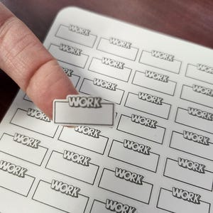 May include: A sheet of white stickers with the word "WORK" printed in a black, bold font. The stickers are rectangular with a tab at the top. A finger is holding one sticker.