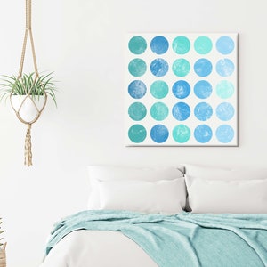 Pode incluir: Cena de quarto com cama branca, manta turquesa e almofadas. Uma tela quadrada com círculos azuis e turquesas está pendurada acima da cama. Uma planta suspensa em um suporte de macramê e uma pequena planta em vaso decoram.