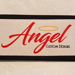 May include: A black canvas wallet with a white background and the word "Angel" in red cursive with a gold halo above the word. The text "Custom Designs" is printed in black below the word "Angel".