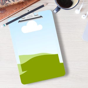 May include: A clipboard with a landscape design featuring a blue sky, white cloud, and green hills. A smartphone, a laptop, a cup of coffee, and a pencil are also on the light wood surface.