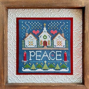 May include: A framed cross-stitch design featuring a blue church with white trim and red hearts, surrounded by small houses and trees. The text "PEACE" is written in white letters below the church. The frame is brown wood with a silver-colored mat.