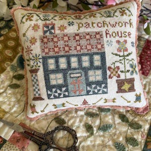 May include: A square pincushion featuring a cross-stitch design of a house with the text "patchwork house" in brown thread. The house is navy blue with a patterned roof, surrounded by floral motifs and small sewing tools, finished with a pink trim.