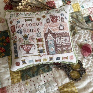May include: A square fabric pincushion featuring a cross-stitch design of a house and the text "hot cocoa house". The embroidery includes motifs of a flower pot, spools of thread, and buttons in shades of brown, blue, and red. The cushion has a light green ruffled trim edge.