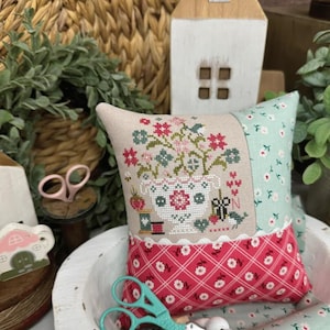 May include: A decorative pillow with a floral cross-stitch design, featuring red and green flowers, a white vase, and a red patterned border. The text "Nettie's Sewing Basket" and "Primrose Cottage" are visible.