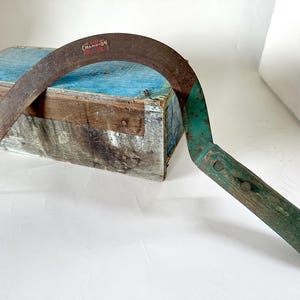 Puede incluir: Una hoz vintage con una hoja curva y oxidada y un mango de madera verde. La hoja está colocada sobre una caja de madera azul y marrón desgastada. La hoz tiene una pequeña etiqueta que dice "Champion".