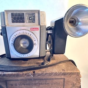 May include: Vintage Kodak Brownie Starmeter camera with a flash attachment. The camera has a silver face with a black and gray body. The flash is black with a large, silver reflector. A black strap is attached to the camera.