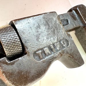 May include: Close-up of a vintage adjustable wrench. The wrench is made of dark metal with the word "TRIO" stamped on the side. The adjustment wheel has a textured, diamond pattern.