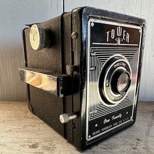 May include: Vintage black Tower camera with silver accents. The camera has the word "TOWER" at the top, and the words "PRECISE" and "One Twenty" are also visible. The camera is marked "SEARS ROEBUCK AND CO USA".