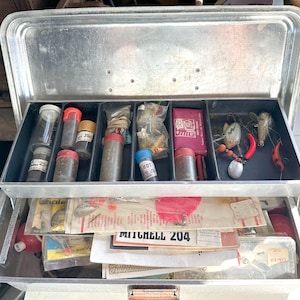 Könnte beinhalten: Geöffnete Metall-Angelbox mit zwei Ebenen. Die obere Ebene enthält transparente Plastikfläschchen mit roten und gelben Kappen, zusammen mit Angelködern und einer rosa Verpackung. Die untere Ebene enthält Angelzubehör und den Text "MITCHELL 204".