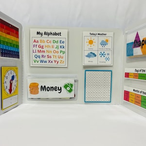 K&ouml;nnte beinhalten: Eine wei&szlig;e Tafel mit bunten Taschen und Beschriftungen. Die Taschen enthalten verschiedene Lernmaterialien, darunter ein Alphabet-Diagramm, ein Wetterdiagramm, ein Hundert-Diagramm, eine Uhr, ein Geld-Diagramm, ein Formen-Diagramm, ein Wochentage-Diagramm und ein Monats-Diagramm.