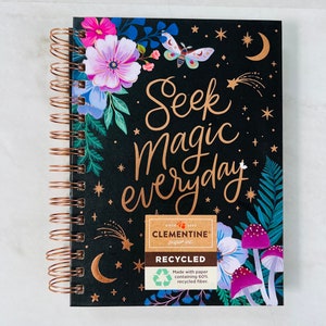 May include: A black spiral bound journal with gold foil lettering that reads "Seek Magic everyday". The cover features a floral design with butterflies, stars, and moons. The journal is made with recycled paper and has a small label that says "Clementine Paper Inc."