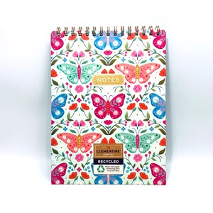 May include: A spiral-bound notebook with a white background and a colorful butterfly pattern. The cover features butterflies in pink, blue, and teal, with floral accents. The word "NOTES" is in gold lettering. The notebook is made with recycled paper.
