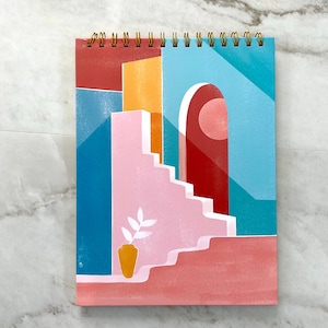 May include: A spiral-bound notepad featuring a colorful abstract illustration of a building with stairs, an archway, and a vase with a plant. The artwork uses shades of red, pink, blue, and orange. The notepad has a gold spiral binding.