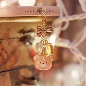 May include: A gold charm with a pink bear, a heart, and a bell hangs from a white phone case. The charm is attached to a gold bow and chain. The phone case is on a wooden surface with a floral design. The background is blurred and features white string lights.