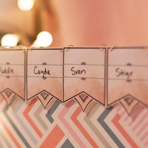 May include: Four beige paper banners with gold clips hanging from a binder. The banners have the names "Puddle", "Cayde", "Sven", and "Stig" written on them. The binder has a geometric pattern in shades of pink, blue, and gray.