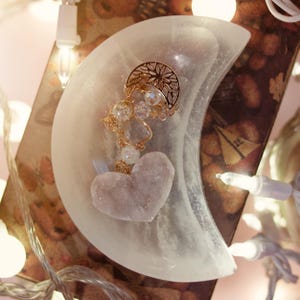 May include: A crescent-shaped, white dish holds a gold-colored charm with clear crystals and a pink heart-shaped stone. The charm is attached to the dish with a delicate gold chain. The dish is illuminated by soft, warm lighting.