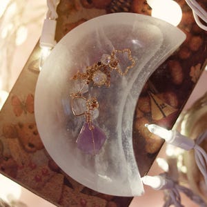 May include: A crescent moon-shaped selenite dish holds a collection of jewelry. The jewelry includes a gold-toned star-shaped charm, a pendant with a purple gemstone, and other delicate pieces. The dish is translucent, and the background is softly lit with fairy lights.